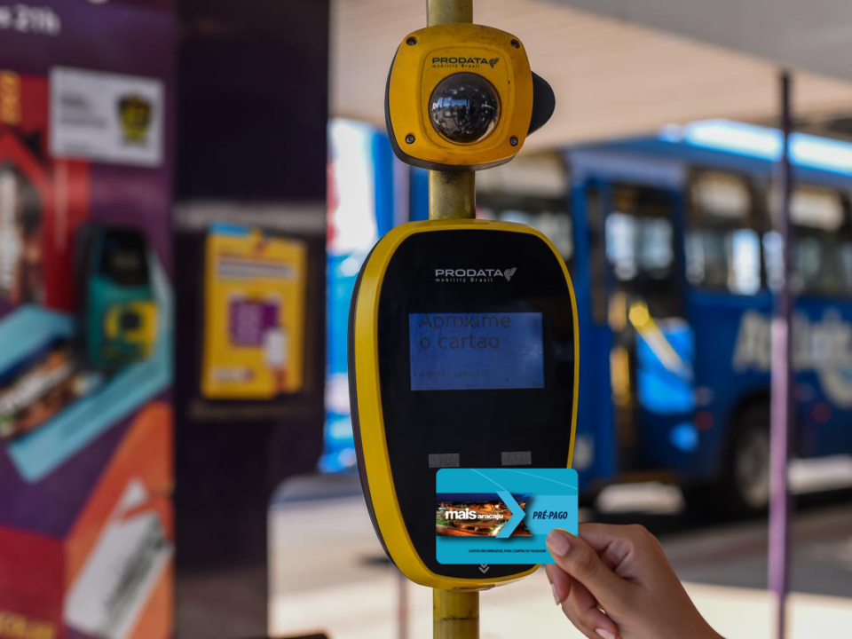 Mão feminina segura cartãos Mais Aracaju Pré-pago próximo a validador de bilhetagem em um terminal de ônibus